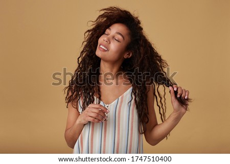 Indoor shot of young positive curly dark skinned brunette lady playing with her hair and smiling gently with closed eyes while posing over beige background