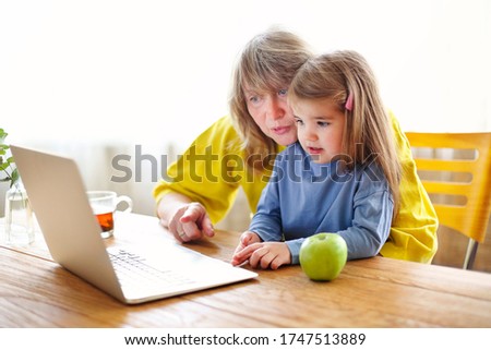 Mature woman and little girl sitting at table and watching cartoon on laptop while spending time at home together