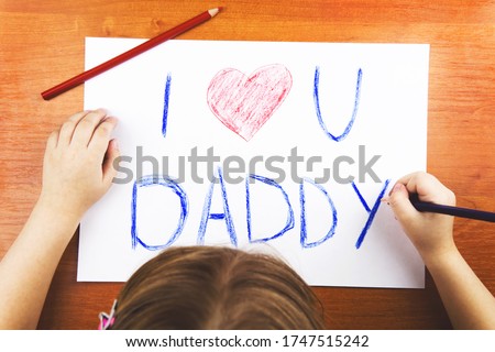 Fathers day concept. Girls hands drawing card with words I Love U Daddy, and red heart, and color pencils on wooden desk.