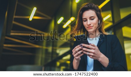 Lifestyle portrait business woman walking in evening city and using mobile gadget, girl banker dressed in business style listen to music wireless earphones and typing text message on smartphone device Royalty-Free Stock Photo #1747522400