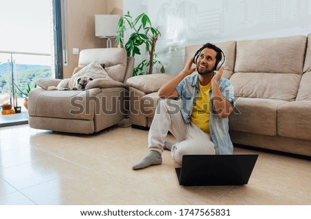 A horizontal shot of a male sitting on the floor in front of a laptop and listening to music