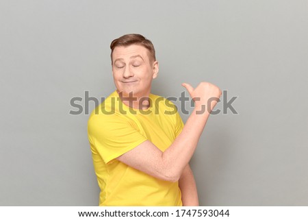 Studio portrait of happy funny blond mature man wearing yellow T-shirt, pointing with thumb at himself, smiling with pleasure, looking successful and proud, standing over gray background