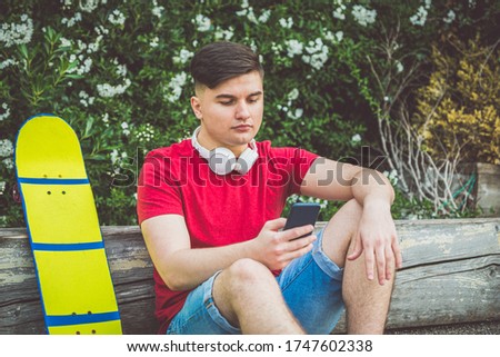 Teen Boy is sitting resting with Longboard - Young adults outdoor skating - A young guy is sitting with a longboard, watching a smartphone in his hands, listening to music with headphones