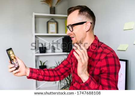 Smiling guy is making a video call via phone, he sits at the workplace and waving hi to his female colleague , a shelf with plants and decor elements on it is on the background