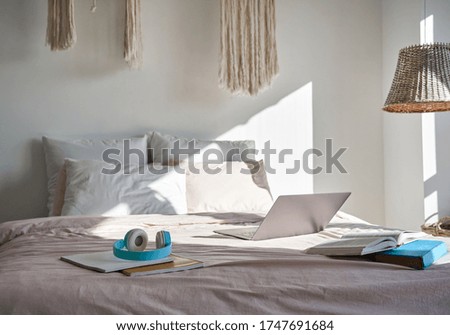Modern bedroom interior with laptop computer, headphones, books on cozy bed in sunny room. Cosy hygge comfortable bed with technology student freelancer workspace at home indoors in morning light.