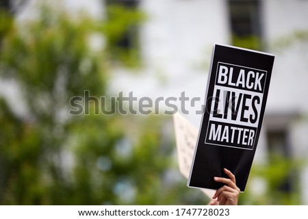 A person holding a printed black sign "black lives matter" at a racially oriented rally. A blurred building and trees in the background Royalty-Free Stock Photo #1747728023