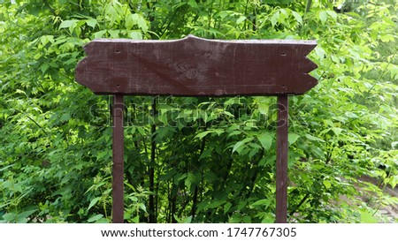 Wooden brown sign on a background of green leaves, bushes and trees in a park or forest. Place for your text or logo, advertisement. Copy space. Blank, blank sign.