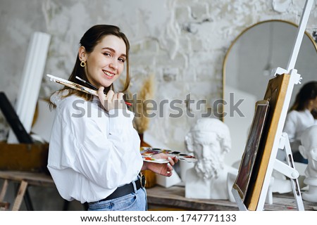  Happy Beautiful Female artist stands  in front of easel in studio, painting a picture on a background of Gypsum  heads.Сreativity, hobby, job and creative occupation concept.Artist 