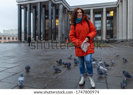 A young woman wearing red jacket and a mask feeding a flock of pigeons in the square near the Russian state library.