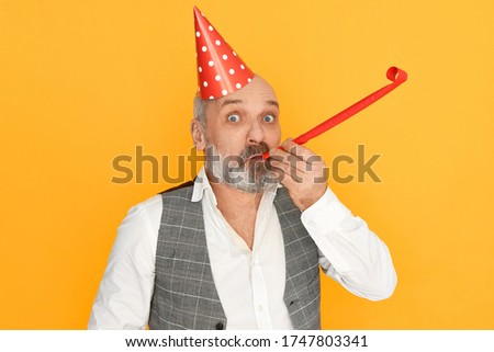 Aging, retirement and celebration concept. Studio image of funny excited senior man with bald head and gray beard wearing elegant clothes and cone hat blowing whistle, celebrating birthday