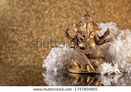Hindu God Ganesha framed by white lilac flowers on a golden bokeh background with reflection