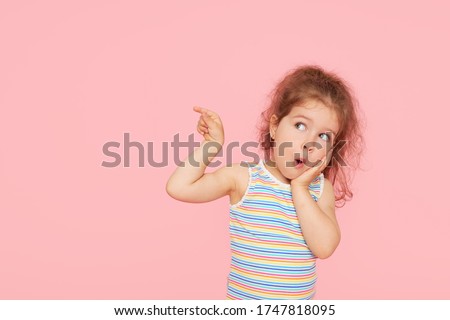 Portrait of surprised cute little toddler girl child over pink background. Looking at camera. Points hands to the left side. Advertising childrens products Royalty-Free Stock Photo #1747818095