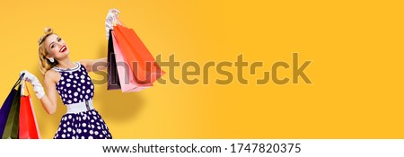Woman in pinup style dress holding, showing or giving many shopping bags, over orange yellow color background with copy space empty area. Big sales or consumer bank credit concept picture. 