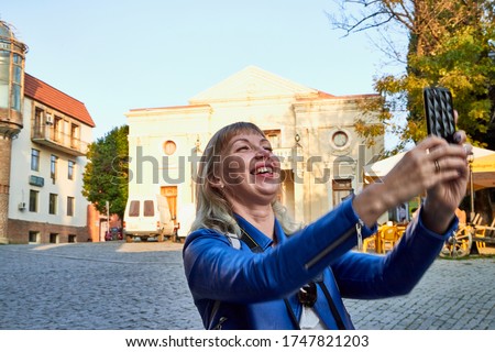 Attractive beautiful young girl holding cell phone in the hands and making selfie. Female photographer traveller walking in city