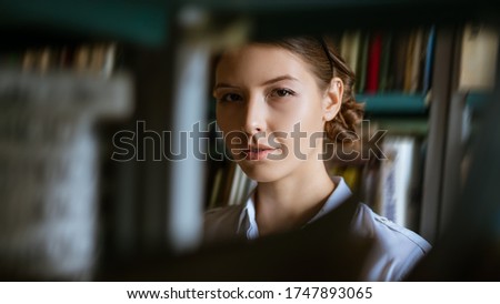 Portrait of a young woman against the background of books in the library, looking through the shelves of books . The concept of preparing for the exams