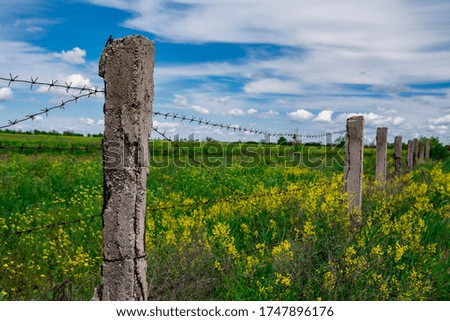 Photo of an old fence, made of  concrete posts connected by rusty barbed wire. A green meadow and a blue sky with a few clouds on the background. 