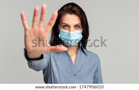 Serious young female in casual wear and surgical mask looking at camera and showing stop gesture while representing concept of social distancing and protection, during coronavirus outbreak