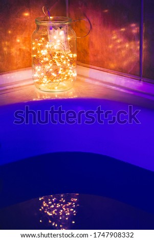 A glass jar with lights stands on the side of a white bath and is reflected in the blue light of the water. House decoration 