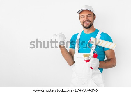 Positive young professional male worker with paintbrush and roller pointing at blank space on white wall and looking at camera with smile