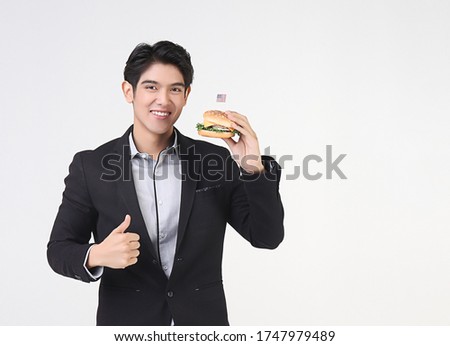 Stylish Asian young and handsome business man wearing dark suit standing on white background , holding hamburger  with American flag,showing his thumb up  and smiling. , independence day concept.