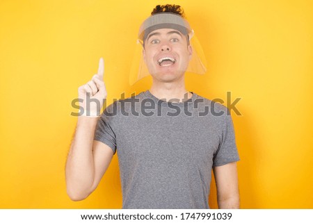 Young caucasian man wearing  t-shirt and face shield and safety gloves  standing over isolated yellow background holding finger up having idea and posing 