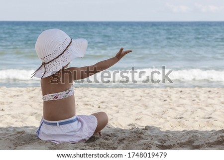 Little girl in a white beach hat and bikini sits on the sand by the sea with her back to the camera, looks at the sea and shows her hand towards the sea