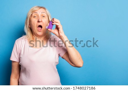 Portrait of an old friendly woman in a casual t-shirt uses an inhaler during an attack on an isolated light background. Emotional face. Asthma, allergy concept