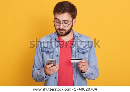 Bearded man paying with credit card on smart phone, looks concentrated, looking at device's screen, holding mobile phone and cardin hands, wearing denim jacket and red shirt, poses against yellow wall