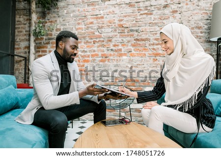Office shot of happy smiling arabic muslim businesswoman satisfied with negotiations results, signing agreement with her handsome African male business partner at office, putting signature on contract