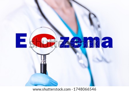A doctor in medical clothes on a light background looks through a magnifying glass at the text ECZEMA. Medical concept.