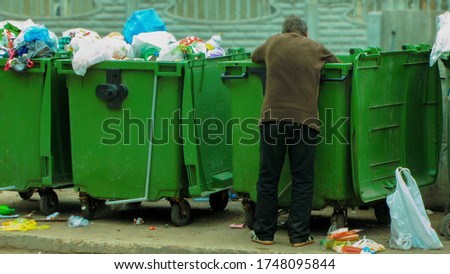 Poor and hungry homeless man in dirty clothes looking for food in the dumpster on the urban street in the city. Social problems of modern society. Lifestyle of tramp, living in the streets. Royalty-Free Stock Photo #1748095844