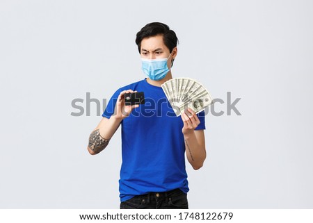 Money, covid-19, easy payment, investment and banking concept. Confused and indecisive asian guy staring frustrated at credit card as holding it and cash, dont understand, wear medical mask