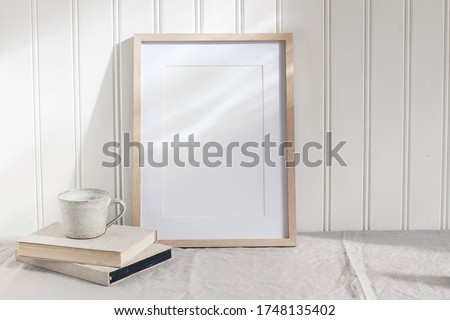 Empty wooden frame mockup on beige linen tablecloth background. White wooden wall paneling background. Artistic still life with cup of coffee and books in sunlight. Scandinavian interior. Art concept.