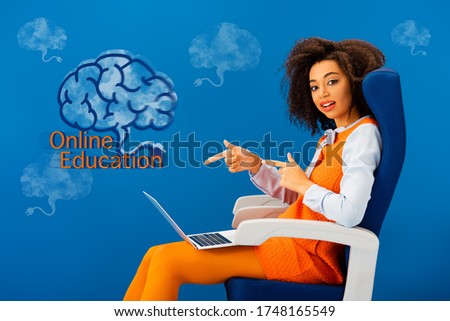 african american woman sitting on seat and pointing with fingers at laptop on blue background with online education illustration
