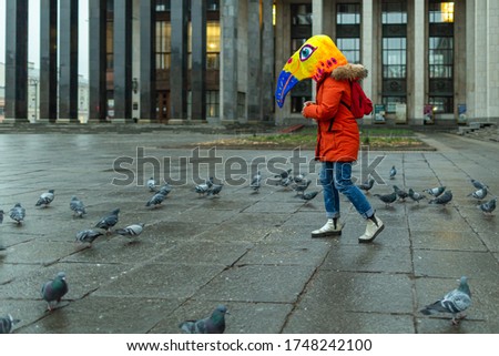 A young woman wearing red jacket and a mask (bird's head) feeds a flock of pigeons in the square near the Russian state library.