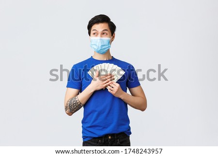 Money, lifestyle, insurance and investment concept. Dreamy happy asian man holding cash and thinking what buy on it, looking away smiling, wear medical mask, grey background