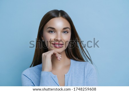 Confident pretty female entrepreneur cares about her complexion, keeps hand under chin, enjoys productive lucky day, listens information carefully, stands against blue background. Sweet cute look