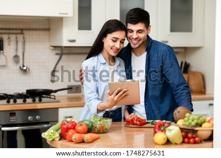 Portrait of lovely young couple using electronic tablet standing in kitchen, hugging and cooking healthy meal