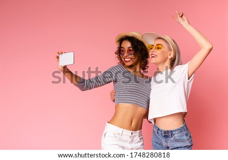 Two cheerful girlfriends in summer slothes taking selfie on smartphone together and showing peace gesture, posing over pink background