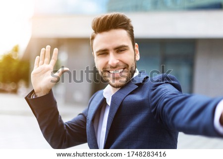 Smiling entrepreneur waving his hand while taking selfie at downtown area