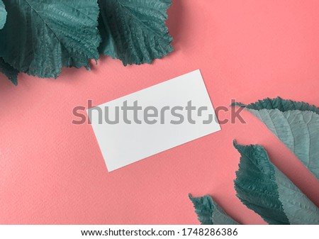 White business card on a soft pink background.There are green leaves in the corners of the photo.You can add text .Photo for placing the company's logo on a business card.