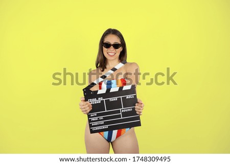 Young beautiful woman standing over yellow isolated background wearing bikini and sunglasses. Very happy and holding a clapperboard