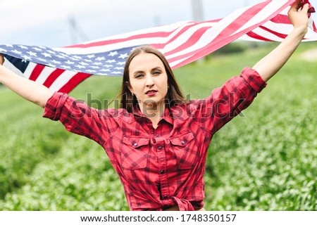 Attractive young woman is walking through green field, her hands raised up with flying US flag