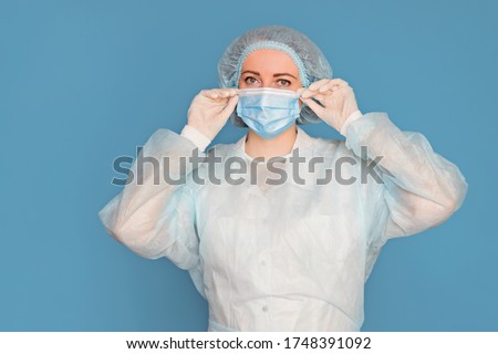 The doctor in a dressing gown, in medical gloves puts on a mask. Surgeon with clinical experience in healthcare, patient care. Copy space, isolated blue background. Royalty-Free Stock Photo #1748391092