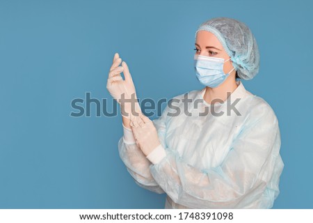Doctor in a dressing gown, mask puts on medical gloves. Surgeon with clinical experience in healthcare, patient care. Copy space, isolated blue background. Royalty-Free Stock Photo #1748391098