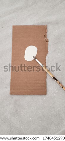 A thin bristled paint brush on a DIY easel made of cardboard. The brush rests in a swatch of white paint.