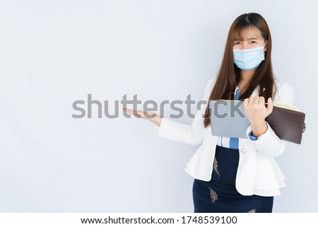 Smiling Asian business woman wearing a medical face mask holding the notebook and pointing hand to side blank space over grey background. Back to the normal concept.