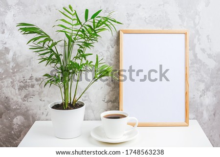Wooden vertical frame with white blank card, cup of coffee and green exotic palm flower in pot on table on gray concrete wall background. Mockup Template for your design, text.