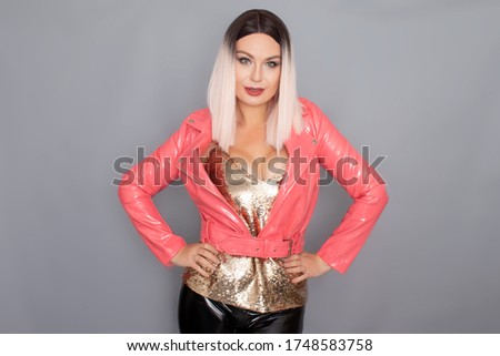 Young stylish blonde woman dressed in a golden top pink and lacquer jacket posing in the studio on a gray background