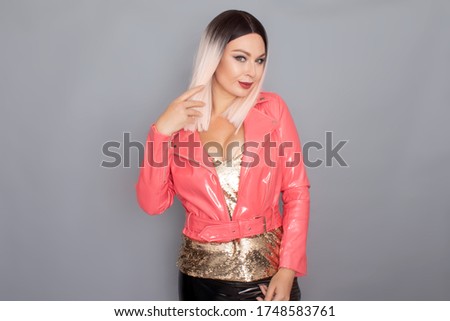 Young stylish blonde woman dressed in a golden top pink and lacquer jacket posing in the studio on a gray background